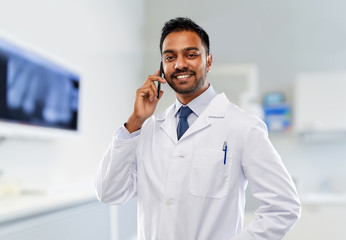 Wall Mural - medicine, dentistry and healthcare concept - smiling indian male dentist in white coat calling on smartphone over dental clinic office background