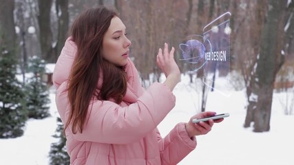 Beautiful young woman in a winter park interacts with HUD hologram with text Web Design. Red-haired girl in warm pink clothes uses the technology of the future mobile screen