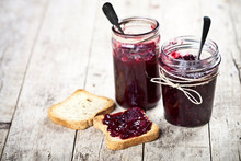 Toasted Cereal Bread Slices And Jars With Homemade Wild Berries  And Cherry Jam And Spoons Closeup On Rustic Wooden Table Background.