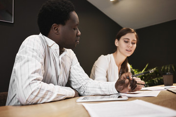 Team of two young professionals European female and African male sharing their ideas and vision while discussing business strategy of common start up project, sitting at desk using digital tablet