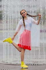 Young wet pretty girl with two braids in yellow boots and with transparent umbrella stands inside of fountain.