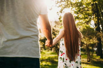 Wall Mural - Back view of a red hair girl leading his boyfriend holding his hand against sunlight walking in the park.