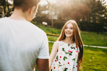 Wall Mural - Portrait of lovely caucasian woman with long red hair and freckles looking at his boyfriend laughing against sunset light while holding hands while dating.