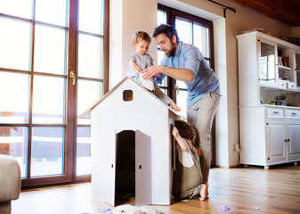 Wall Mural - Two toddler children with father playing with paper house indoors at home.