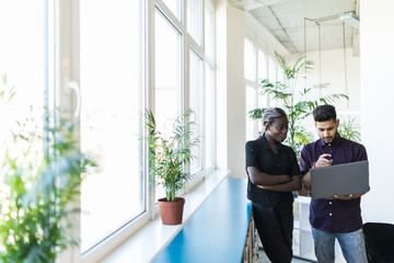 Indian Businessman and african businesswoman using a laptop together while standing in front of office windows overlooking the city