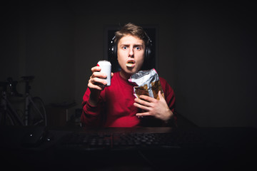 Wall Mural - Young man sitting at the table in the room eats chips and holds drink in his hands, looks at the computer screen,astonished face.Surprised teen eats snack, watches a scary video on a computer at night