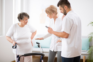 Wall Mural - Grey senior lady with walker during physiotherapy with professional female doctor and male nurse