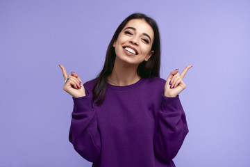 Pretty adult girl with broad smile, wears purple sweatshirt, pointing fingers up at copyspace, isolated over violet wall