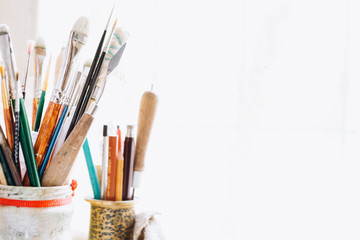 Artist's tools, brushes, paints and a palette lie on a old white wooden sills near light window background. Copy space for text