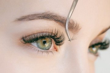 Eyelash Extension Procedure. Close up view of beautiful green female eye with long eyelashes. Stylist holding tweezers, tongs and making lengthening lashes for girl in a beauty salon. Beauty Concept.