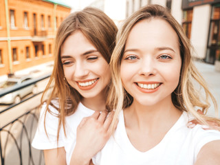 Two young smiling hipster blond women in summer white t-shirt clothes. Girls taking selfie self portrait photos on smartphone.Models posing on street background.Female showing positive face emotions