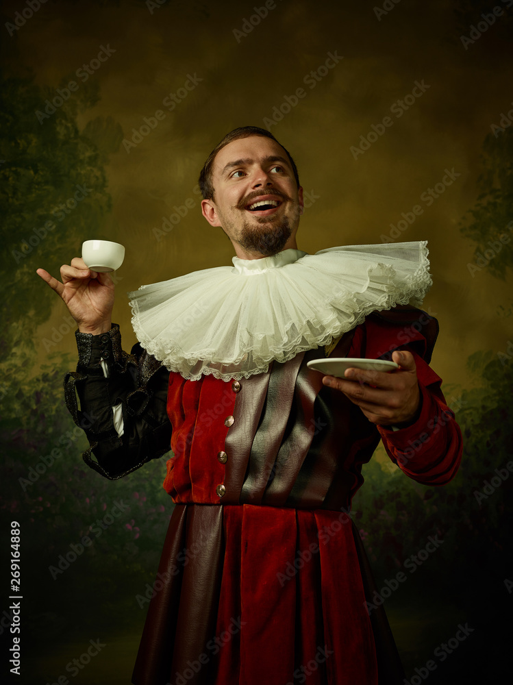 Young man as a medieval knight on dark studio background. Portrait in low key of male model in retro costume. Drinking morning coffee. Human emotions, comparison of eras and facial expressions concept - obrazy, fototapety, plakaty 
