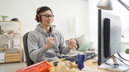 Wall Mural - asian male gamer with headphones emotionally rejoices in victory looks at computer screen. joyful young man university student on summer break vacation stay at home playing video games eating snack.