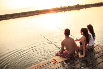 Group of friends sitting on a pier by the lake and fishing. They're joying in beautiful summer sunset.	