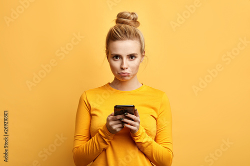 Fotografía angry sad woman with hairbun annoyed by something while using phone, girl has received bad sms, text message isolated yellow background