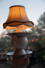 Table lamp made from old crockery teapot, plates and cups with vintage lampshade on top. Retro style lamp on blurred background through window. Reusing old kitchen utensil as interior lighting decor