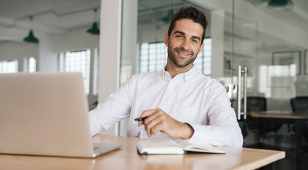 Smiling businessman writing down notes while working on a laptop