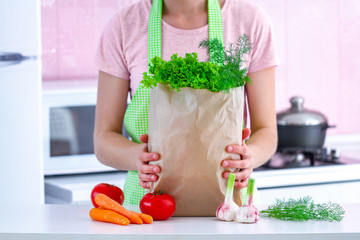 Cooking woman in apron holding a craft paper bag full of fresh organic vegetables at kitchen. Healthy food and balanced diet