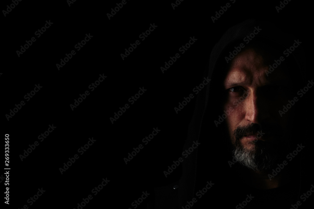 Brutal man with a gray beard in the hood with sharp shadows on a black background. Copy space. - obrazy, fototapety, plakaty 
