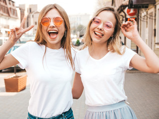 Portrait of two young beautiful blond smiling hipster girls in trendy summer white t-shirt clothes. Sexy carefree women posing on street background. Positive models having fun.Shows peace sign