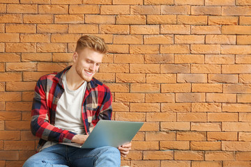 Wall Mural - Handsome young man with laptop sitting near brick wall