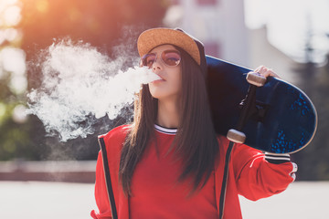 Vaping girl. Young woman with skateboard vape e-cig. Pretty young female in black hat, red clothing vape ecig, vaping device at the sunset. Toned image. Hip-hop style.