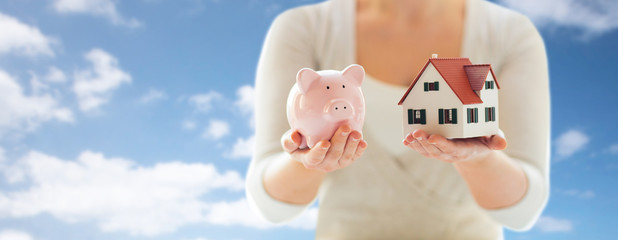 Poster - mortgage, investment, real estate and property concept - close up of woman holding home or house model and piggy bank over blue sky and clouds background