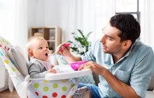 Family, Food, Eating And People Concept - Happy Father Feeding Little Baby Daughter Sitting In Highchair With Puree By Spoon At Home