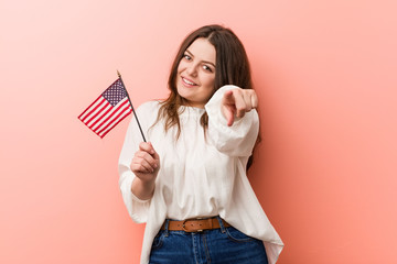 Wall Mural - Young curvy plus size woman holding a united states flag cheerful smiles pointing to front.