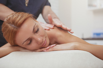 Close up of a beautiful red haired woman relaxing at spa center, receiving full body massage, copy space. Stunning woman smiling with her eyes closed, getting soothing massage