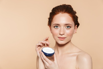 Poster - Amazing young redhead woman posing isolated over beige wall background take care of her skin with cream.