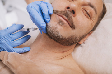 Handsome cheerful bearded mature man getting face filler injections by professional cosmetologist. Close up cropped shot of beautician doing anti-wrinkle treatment for male client