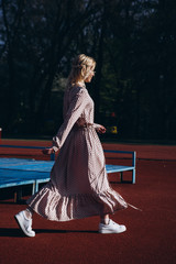 Stylish modern woman with light hair in a light summer dress under sunlight poses for a photo on a sports ground in the summer.