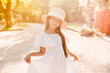 © Look! - Outdoor portrait of cute fashionable girl wears boater decorated with ribbon, playing with her dress. Charming little lady in straw hat dancing in park in sunny day.