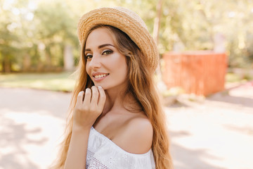 Wall Mural - Close-up portrait of fascinating young woman playfully posing in sunny day standing in park. Outdoor photo of gorgeous girl with beautiful light-green eyes smiling, touching her chin.