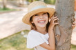 © Look! - Laughing little girl with lightly-tanned skin posing in park touching tree. Outdoor close-up portrait of cheerful dark-haired kid in vintage hat with ribbon having fun in garden.