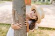 © Look! - Funny dark-haired kid with big eyes and smile embracing tree in park. Outdoor portrait of happy little girl in straw hat enjoying summer vacation.