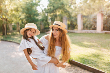 Poster - Inspired woman in good mood holding daughter and laughing while posing in alley with green trees. Outdoor portrait of adorable young mom carrying girl in white dress with cute smile.