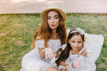 Poster - Stunning young woman and pretty girl sharing love with world posing for photo on picnic. Two amazing sisters in similar white attires sending air kisses relaxing on the grass in morning.