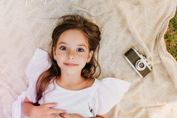 Wall Mural - Amazing dark-eyed girl lying on blanket with surprised smile beside camera. Overhead outdoor portrait of little lady in white dress relaxing on the grass in park.