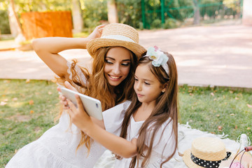 Smiling adorable woman with trendy make-up posing while her daughter holding white smartphone. Outdoor portrait of pretty young mother and her child in white dress making selfie in park.