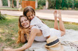© Look! - Funny young woman fooling around with daughter lying on white blanket and making faces. Outdoor portrait of pretty girl sitting on mom's back and embracing her.