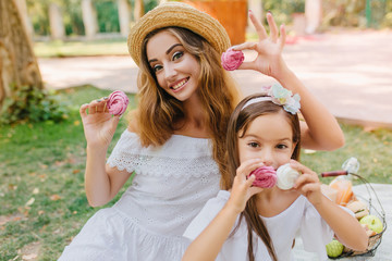 Outdoor portrait of cheerful young woman in vintage dress and joyful girl with ribbon in dark hair posing on nature background. Happy mother and daughter holding tasty cookies in park.