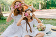 © Look! - Stylish young woman came with pretty daughter to park to spend weekend together. Outdoor portrait of brown-haired girl joking with mother while eating cookies on blanket.