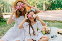 Stylish Young Woman Came With Pretty Daughter To Park To Spend Weekend Together. Outdoor Portrait Of Brown-haired Girl Joking With Mother While Eating Cookies On Blanket.