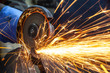 Close-up on the sides fly bright sparks from the angle grinder machine. A young male welder in a white working gloves grinds a metal product with angle grinder in the garage