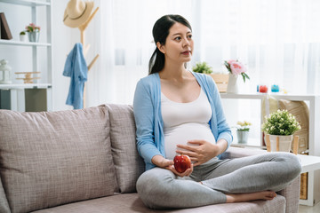 Wall Mural - Pregnant beautiful asian girl hold red apple. Attractive mother feels relaxed and happy. charming woman has good health takes good care of herself by eating healthy food sitting on couch sofa at home