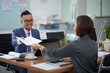 © DragonImages - Smiling Asian businessman in eyeglasses sitting at the table in front of laptop computer and giving some documents to his colleague opposite him at office