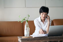 Asian Self Employed Woman Sitting On Couch  Talking On Phone & Working On Laptop At Home