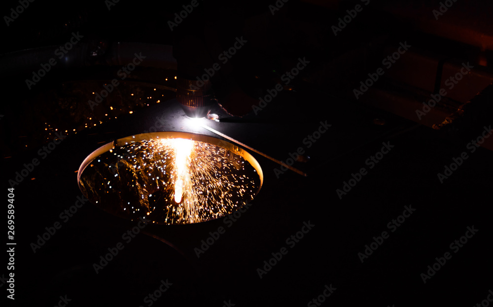 An automatic plasma cutter in a workshop cutting out a circle in a sheet of metal, dark scene, illuminated by sparks and flame.